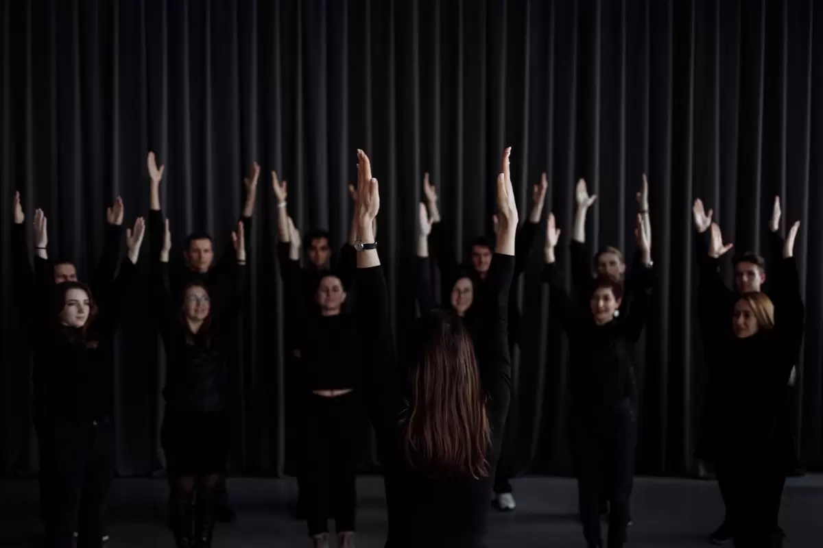 A female conductor has her back to us as she faces the choir while leading them in a warm-up exercise. Everyone has both hands high in the air. All are dressed in black.