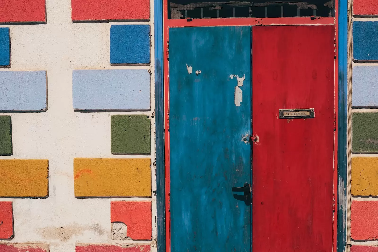 Closed double door painted blue on the left and red on the right, in a cement wall with embedded orange-pink, gold, green and light blue painted bricks.