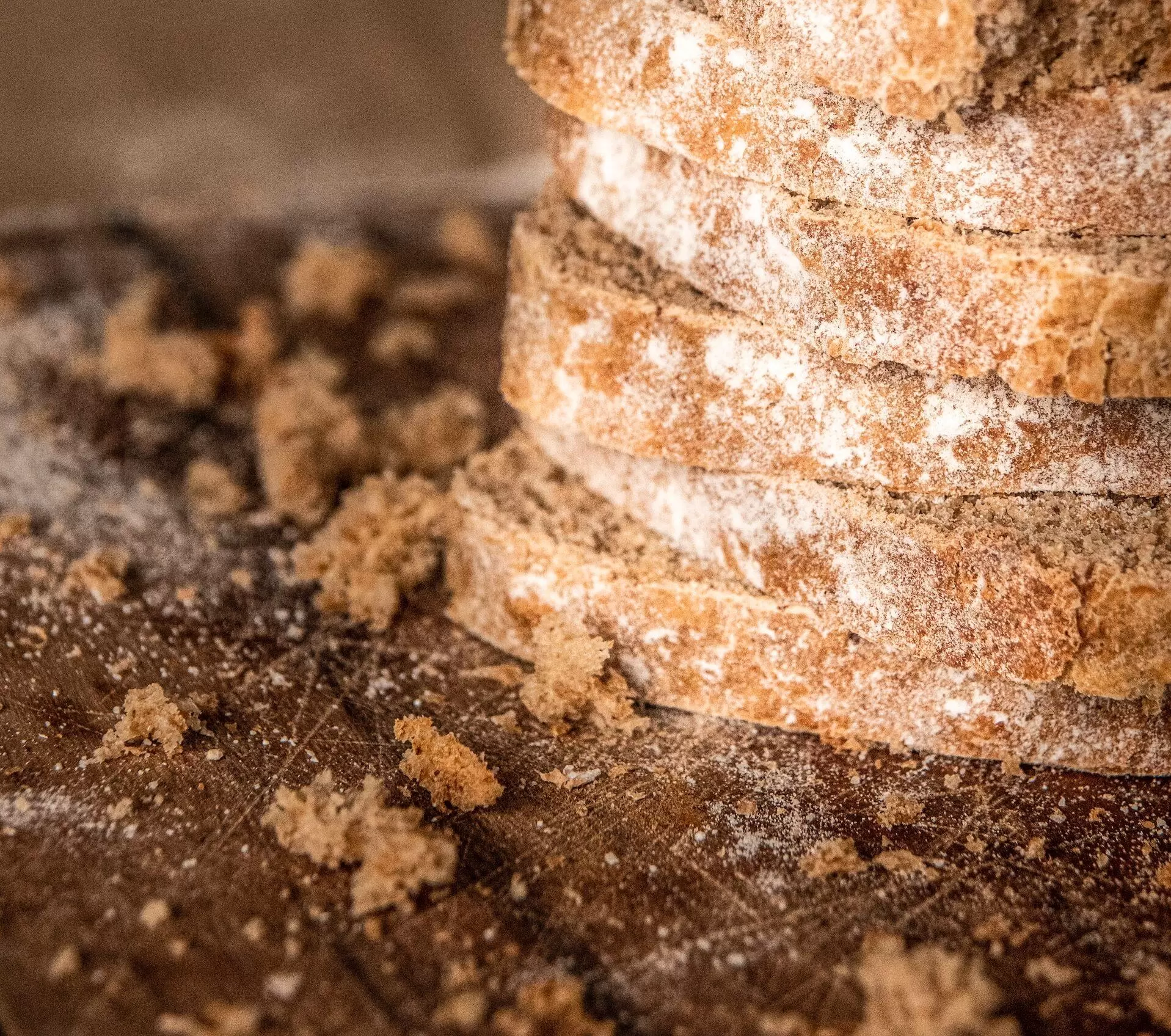 Bread crumbs rest on a wooden table beside a stack of thick, whole-grain bread slices.